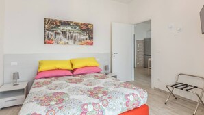 Une chambre à coucher avec un lit, des oreillers, une table de chevet, une lampe, un tableau accroché au mur et une salle de bain visible à travers une porte ouverte.