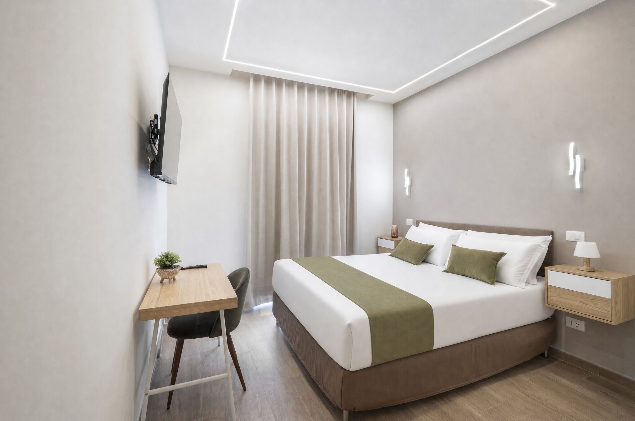 A modern hotel room with a bed, bedside table, lamp, and a desk with a chair.