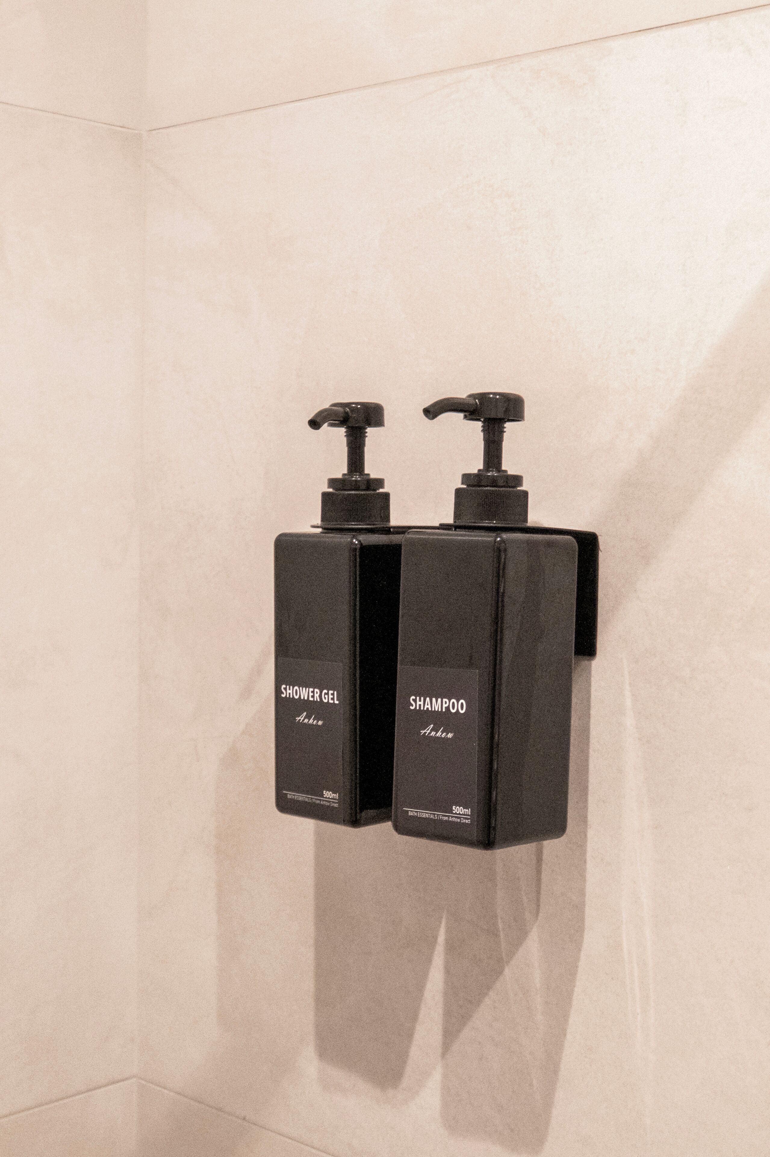 Two black dispensers labeled 'Shower Gel' and 'Shampoo' mounted on a wall.