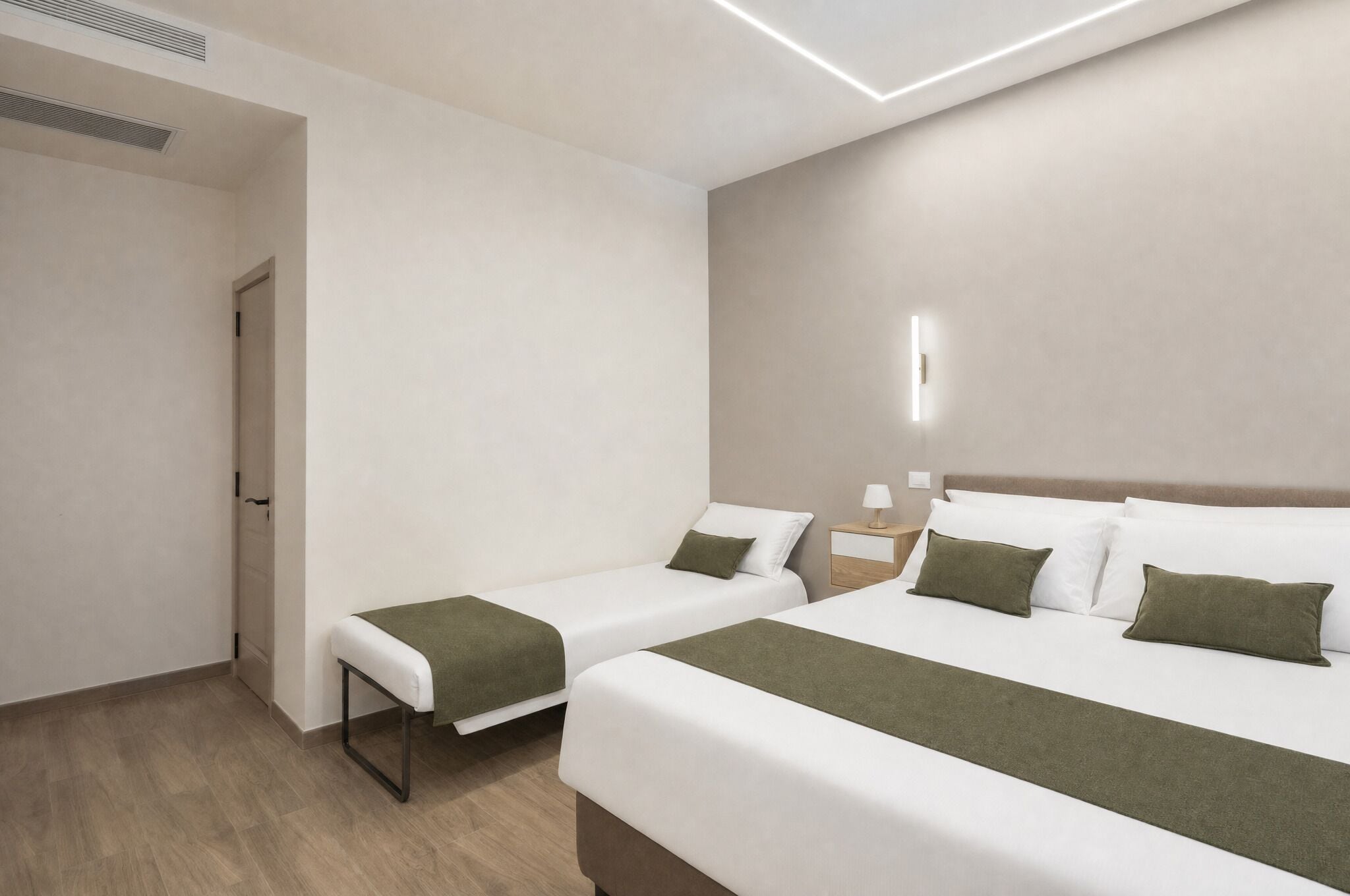 A hotel room with two beds, a bedside table with a lamp, and a bench.