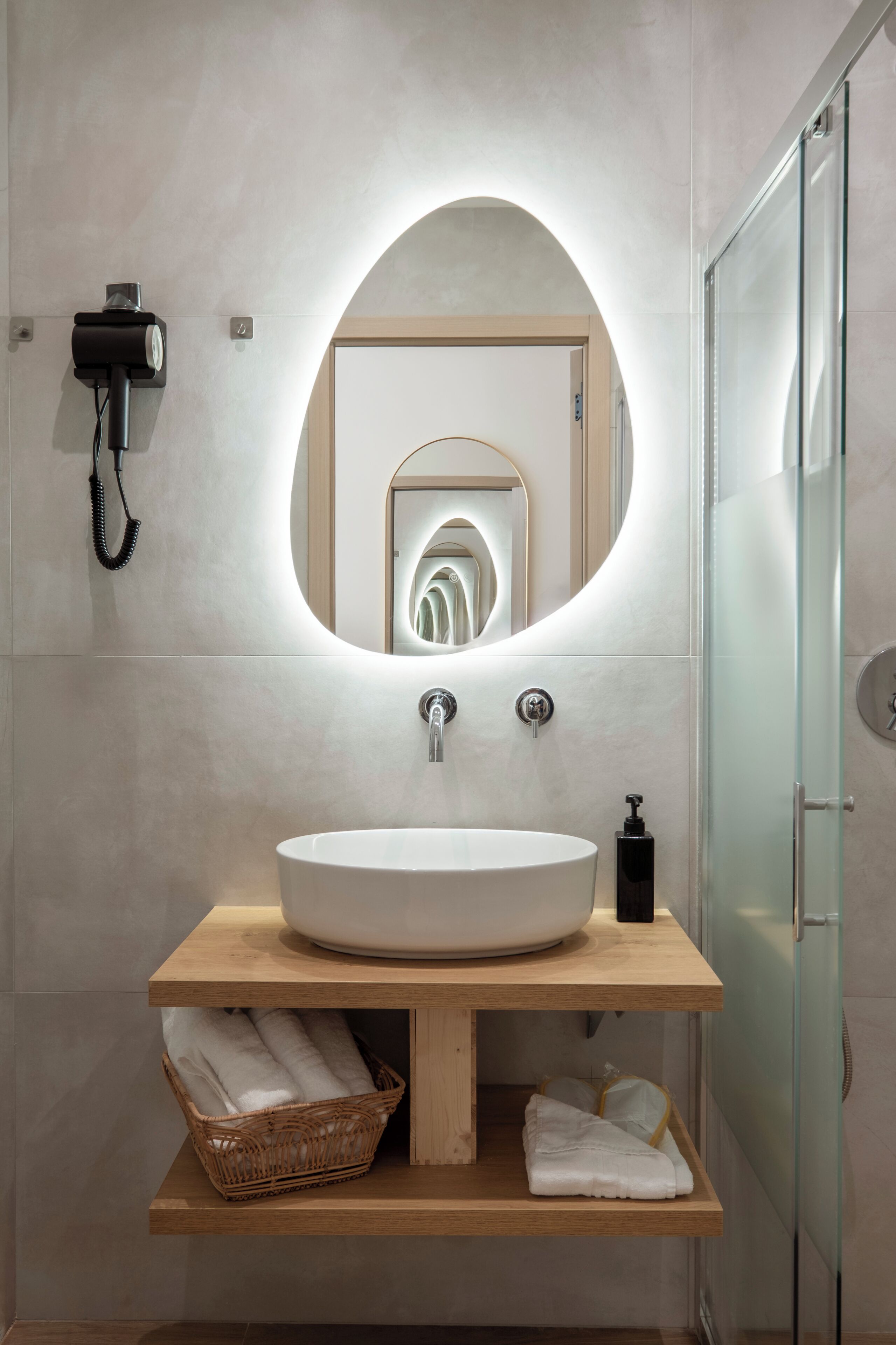 Une salle de bain moderne avec un grand miroir ovale, un meuble-lavabo en bois, une cabine de douche en verre et un panier en osier.