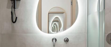 Une salle de bain moderne avec un grand miroir ovale, un meuble-lavabo en bois, une cabine de douche en verre et un panier en osier.