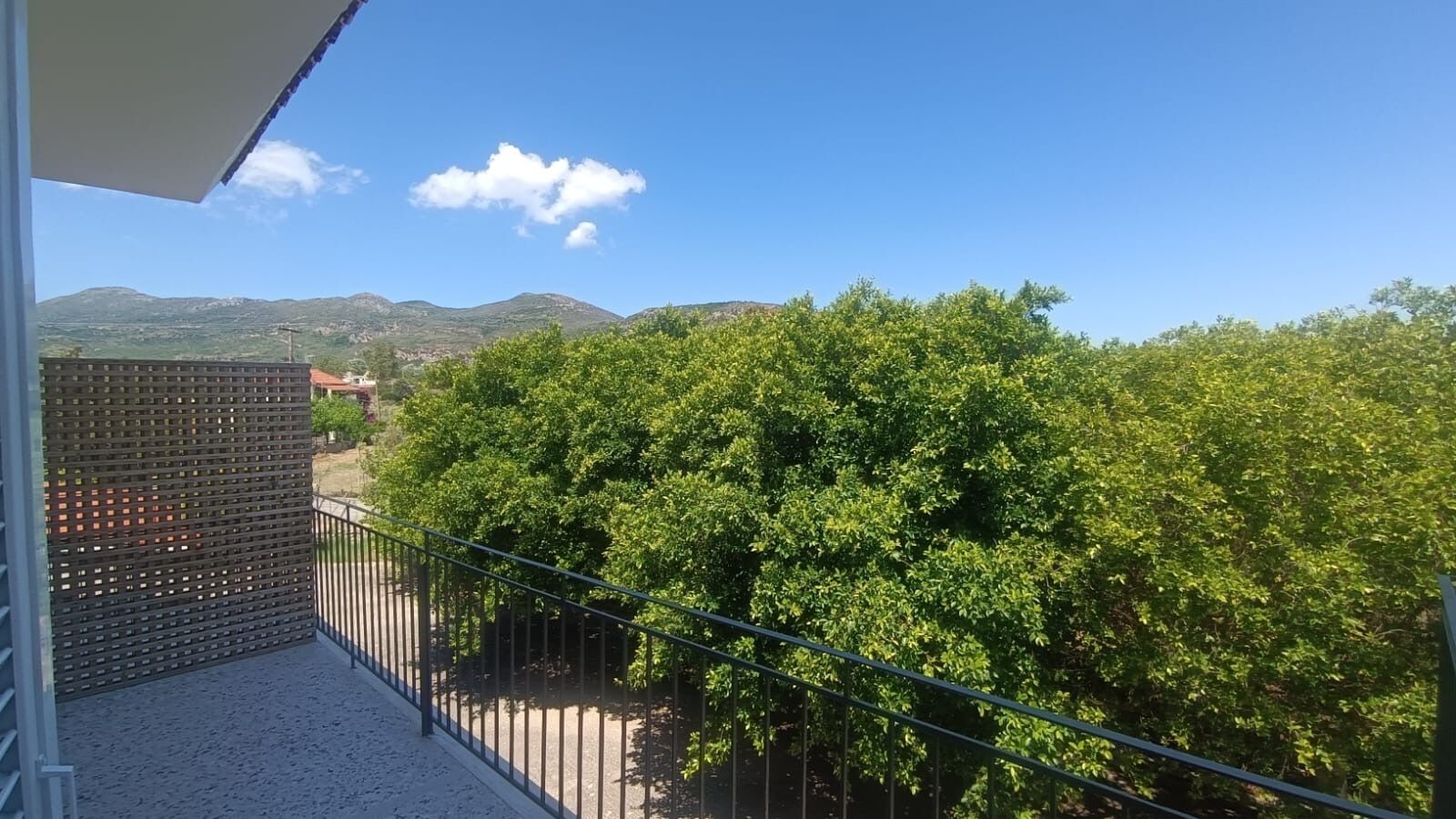 A balcony with a metal railing, a view of green trees, and distant mountains under a clear sky.