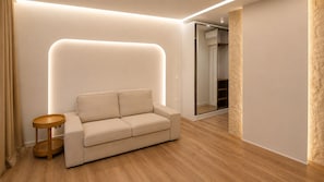 A modern room with a beige sofa, a small wooden side table, and a glass door leading to a bathroom. - Elysium Apartments (Kefalonia)