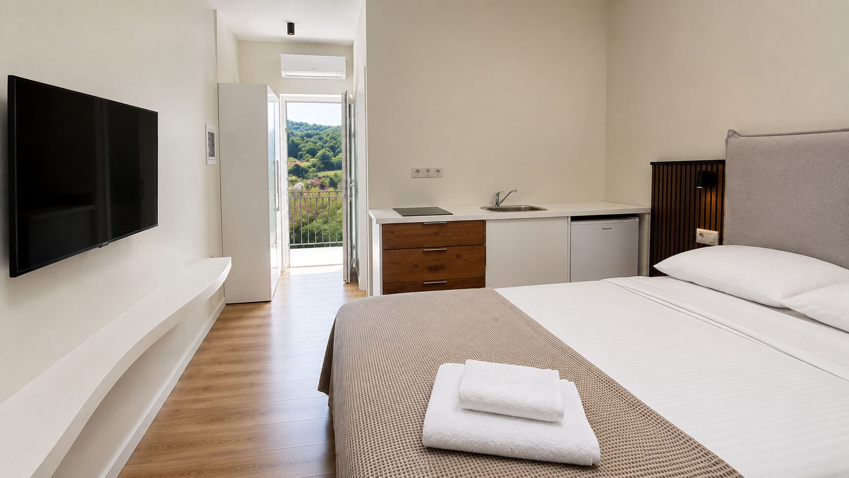 Un dormitorio con televisor, cama con ropa blanca, baño con lavamanos y vistas a zonas verdes.