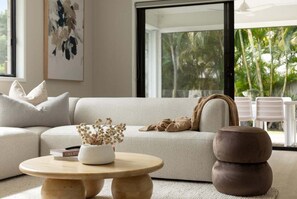 A modern living room with a sofa, a round wooden coffee table, and a view of tropical trees through large windows. - Blue Gum Hideaway - Pets by approval (Sunshine Coast)