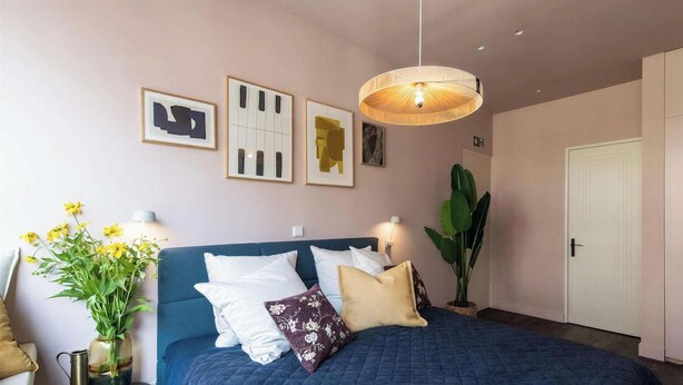 Een slaapkamer met een bed, een nachtkastje met een lamp, een grote potplant en kunst aan de muur.