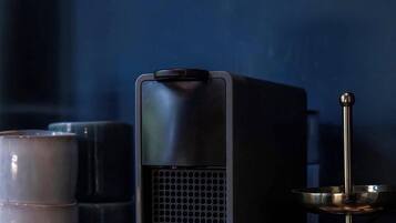 A black speaker, a KRUPS coffee grinder, and a candle holder with candles on a dark surface.