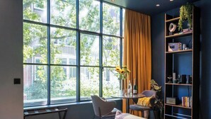 Een kamer met grote ramen, een boekenkast, een tafel met een vaas zonnebloemen en een stoel.