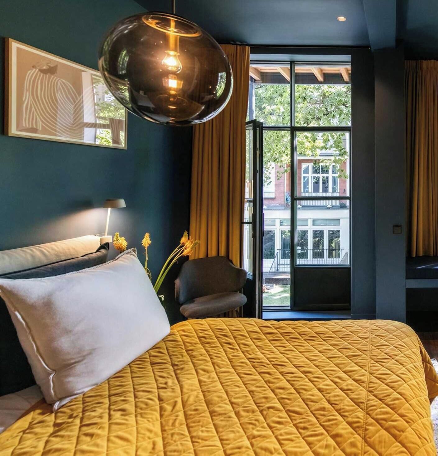 Une chambre avec un grand lit, un luminaire suspendu, une chaise et une vue sur l’extérieur.