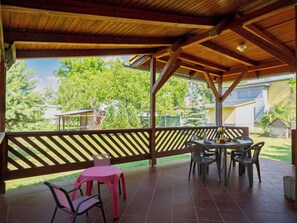 Un patio cubierto con una mesa y sillas, techo de madera y vista a una zona residencial con árboles y casas.