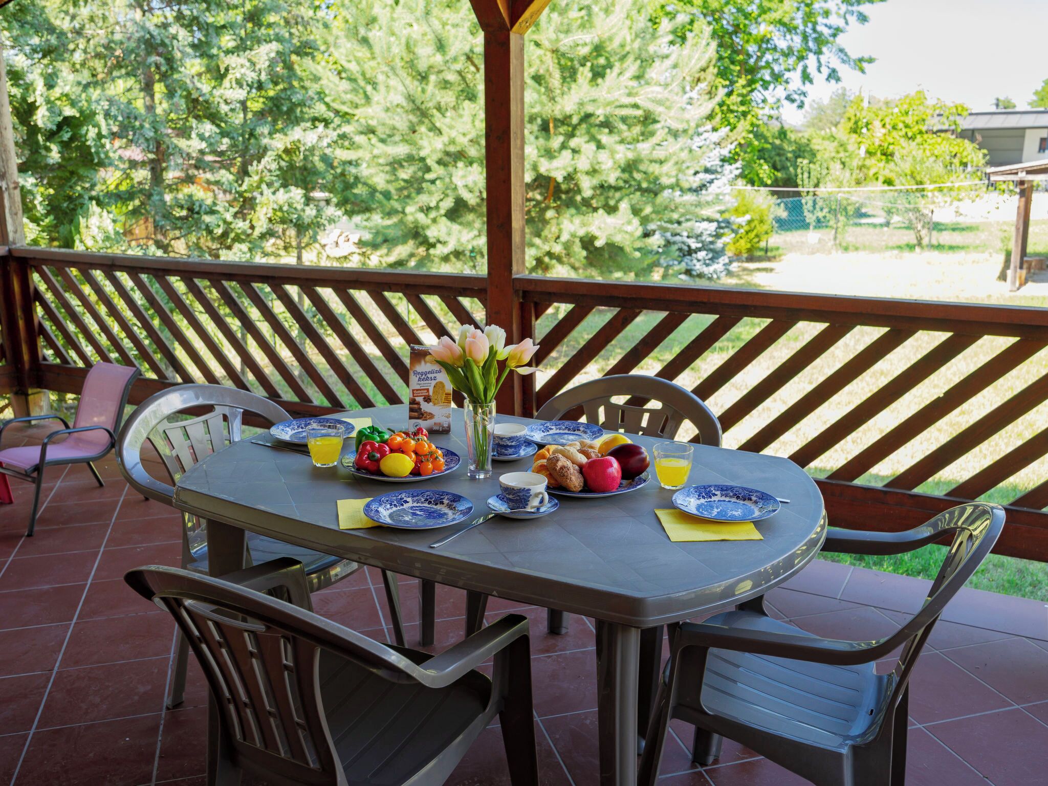 Un patio al aire libre con una mesa lista para el desayuno, que incluye platos, tazas y un florero con flores.