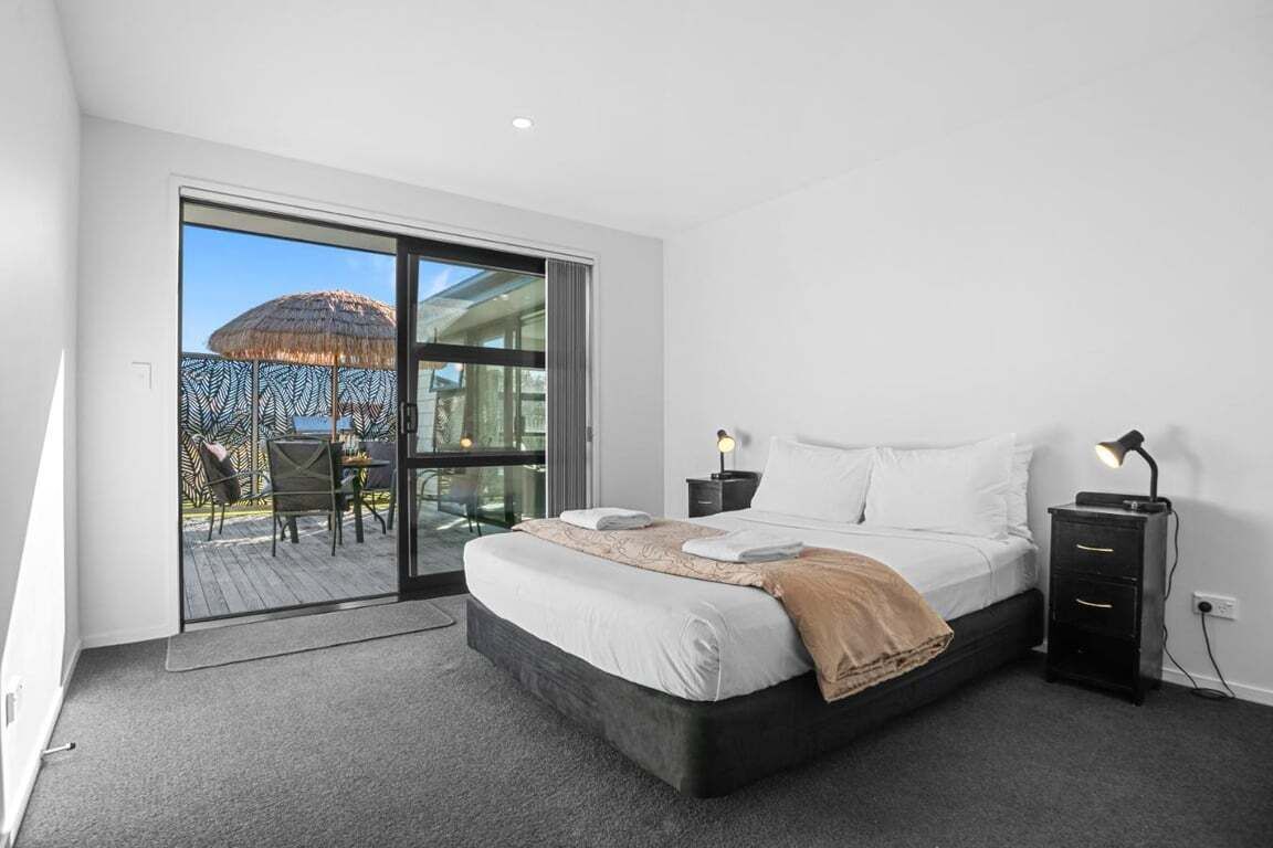 A bedroom with a large bed, a nightstand, a desk, and a view of the outdoors.