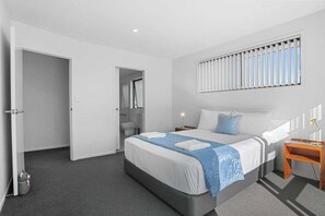 Ein modernes Hotelzimmer mit einem großen Bett, einem Nachttisch, einer Fenster mit Jalousien und einer Tür zu einem Badezimmer.