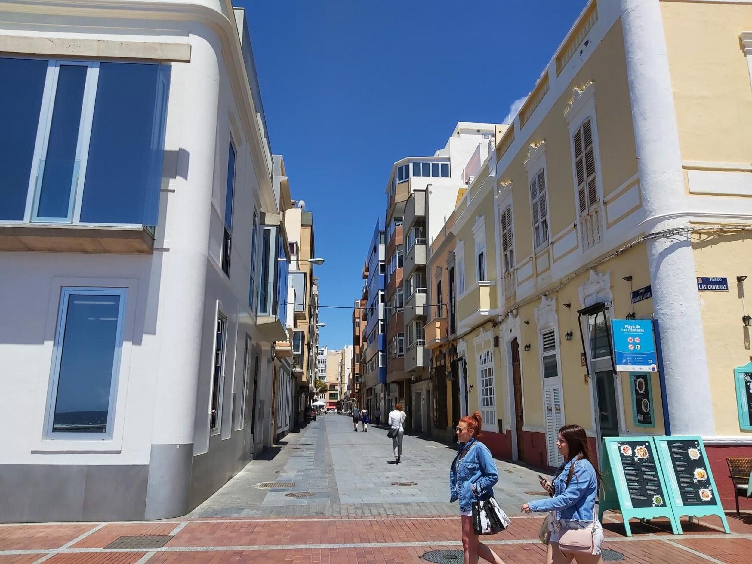 A narrow street lined with multi-story buildings, a clear blue sky, and a few pedestrians.