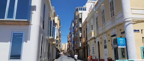 A narrow street lined with multi-story buildings, a clear blue sky, and a few pedestrians.