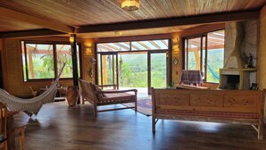 A spacious room with wooden floors, a hammock, a sofa, a fireplace, and large windows offering a view of greenery.