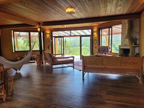 Habitació àmplia amb terres de fusta, un hamaca, un sofà, una llar de foc i grans finestres que donen vistes a vegetació.