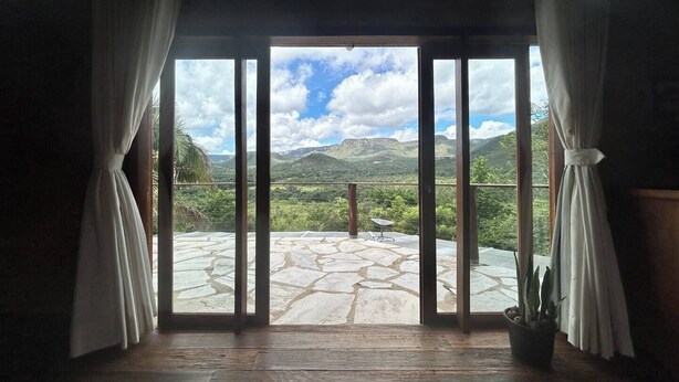 A room with a large glass door leading to a balcony with a stone floor and a view of green hills.