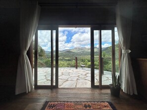 Habitació amb una gran porta de vidre que dona a un balcó amb sòl de pedra i vista a turons verds.