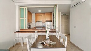 A dining area with a wooden table, white chairs, a vase with white flowers, and a magazine. A kitchen is visible in the background with wooden cabinets and a refrigerator. - Vacation Apartment 'Balcón De Los Gigantes' with Sea View, Wi-Fi and Air Conditioning (Puerto de Santiago)