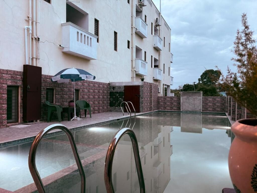 Uma piscina com barreira de vidro, um edifício com varandas e mobiliário de exterior.