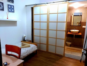 Ein kleines Zimmer mit einem Bett, einem Sessel, einem Tisch, einer Schiebetür, einem Waschbecken und einem Spiegel.