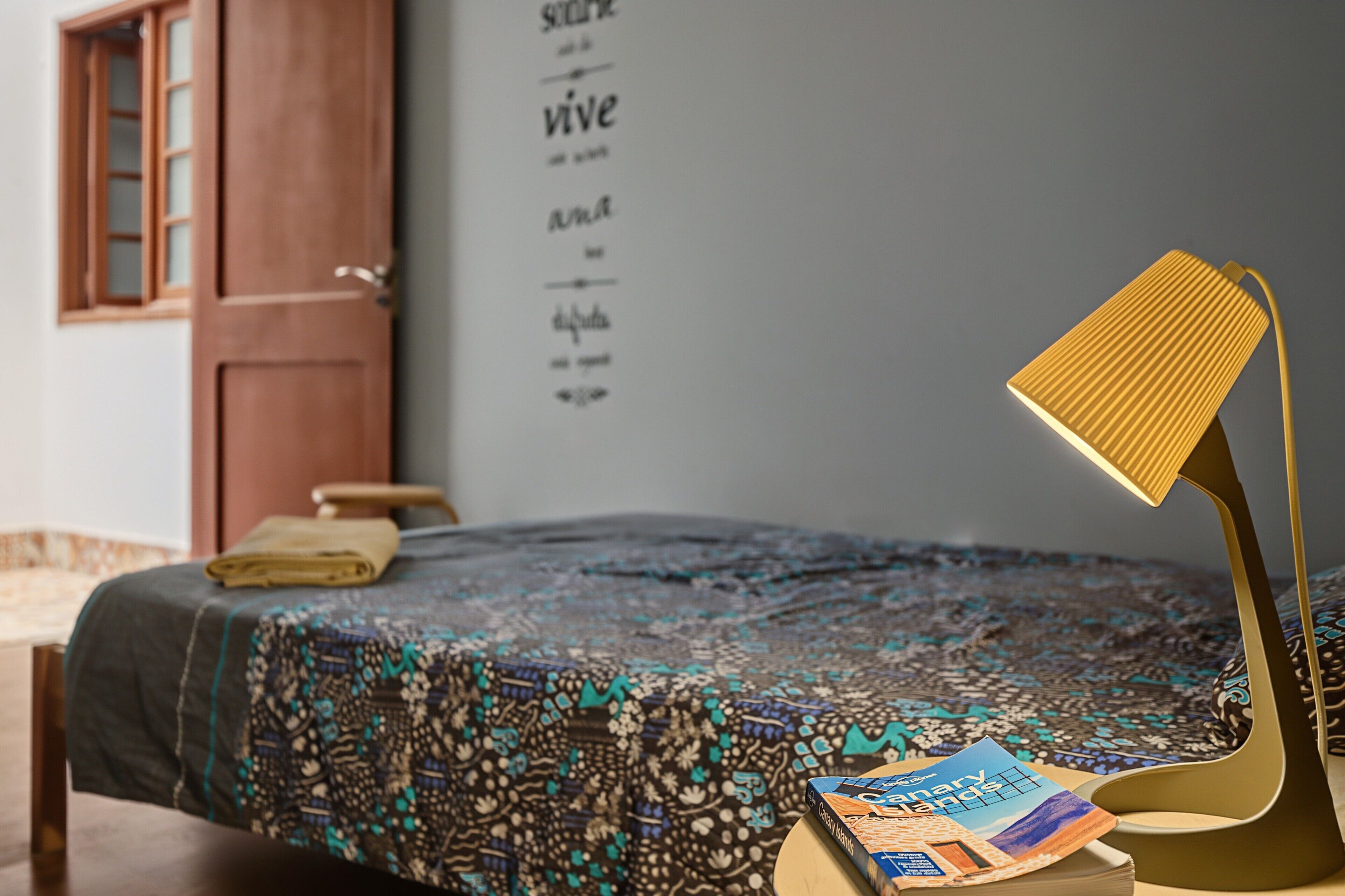 A bed with a patterned bedspread, a lamp, and a book on a bedside table in a room with a wooden door and a wall with motivational quotes.