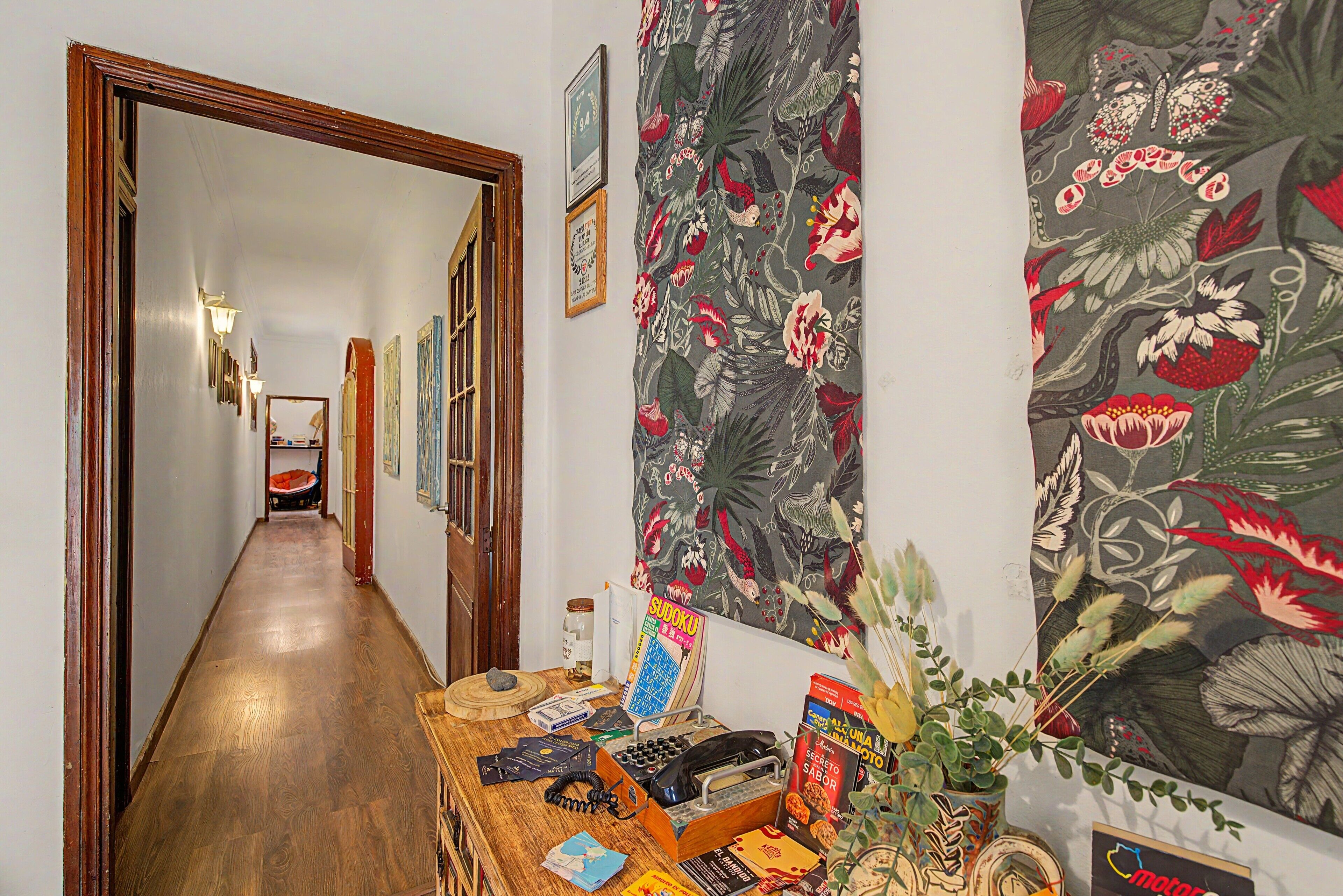A hallway with a wooden floor, a desk with various items, and a wall with floral wallpaper.