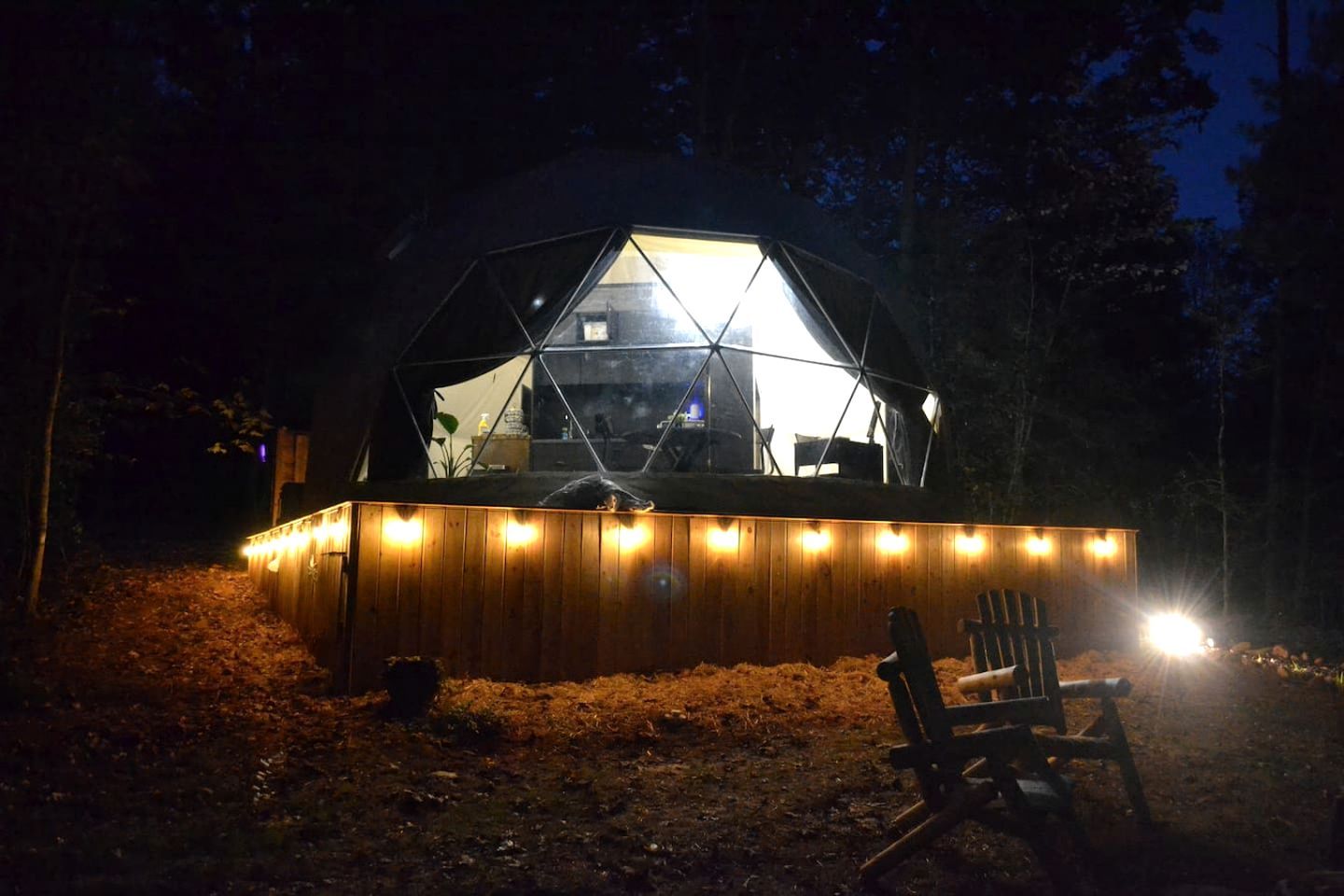 Una estructura de domo geodésico con una terraza de madera y una silla, rodeada de árboles.