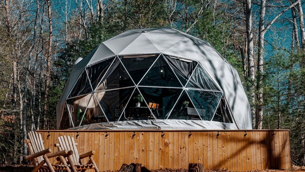 A geodesic dome structure with a wooden deck and chairs in a forest setting.