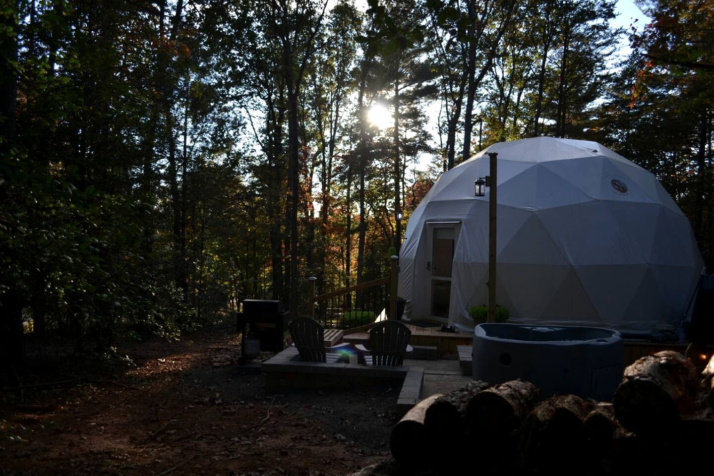 A geodesic dome structure in a forested area with a deck, outdoor seating, and a fire pit.