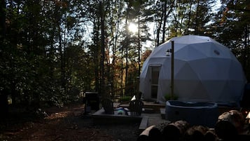 A geodesic dome structure in a forested area with a deck, outdoor seating, and a fire pit.