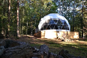 Une structure en forme de dôme géodésique située dans une zone boisée, avec une terrasse en bois et des sièges.