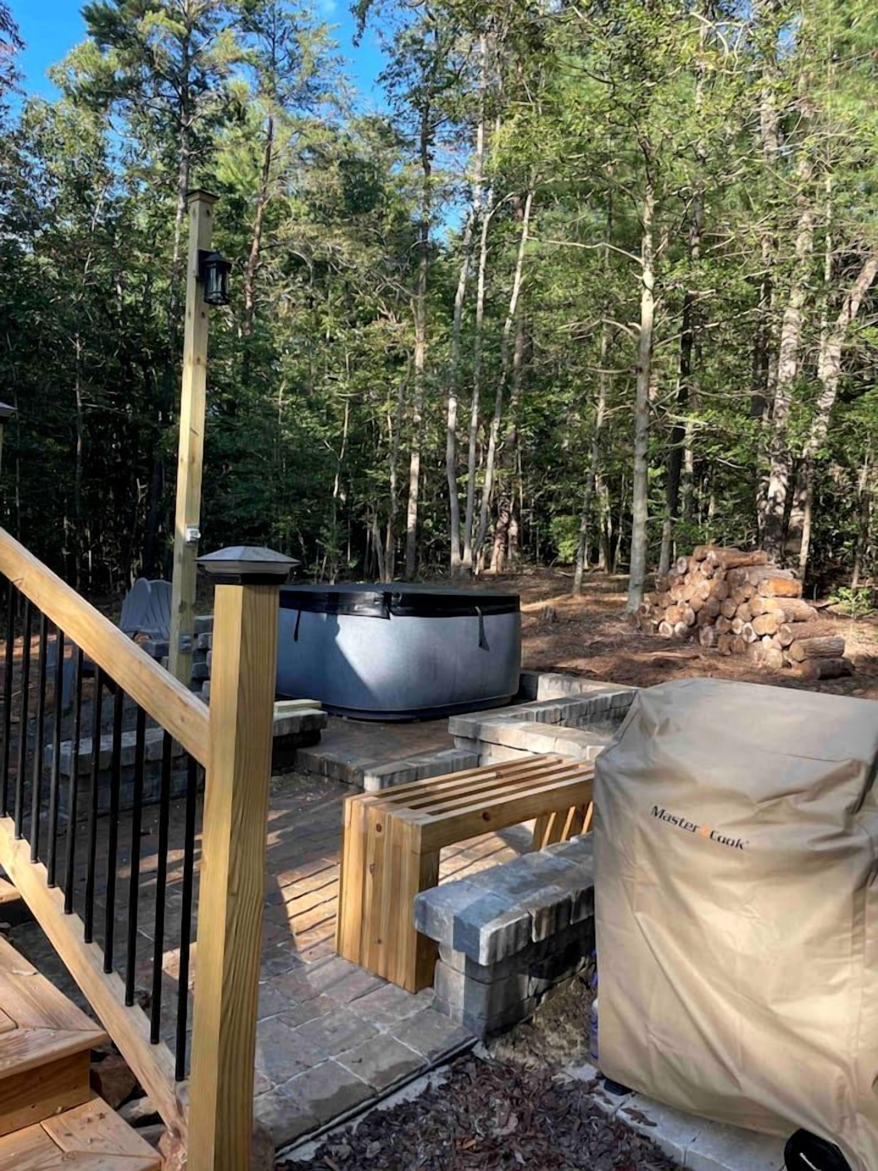 Una terraza con un jacuzzi, una parrilla cubierta y una escalera de madera que lleva a un área de terraza rodeada de árboles.