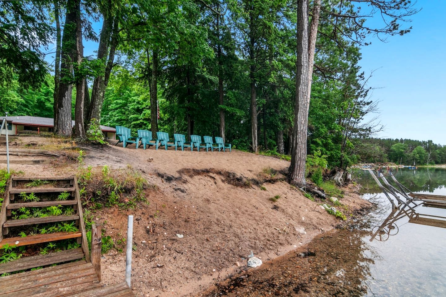 A sandy beach area with wooden steps leading down to the water, surrounded by trees and a row of green chairs.
