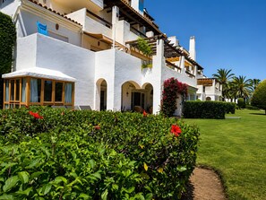 A white building with multiple balconies, a covered entrance, and a well-maintained garden with green hedges and red flowers. - Apartment 'Beachside  San-Pedro' with Mountain View, Wi-Fi and Air Conditioning (Marbella)
