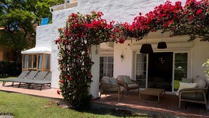 Ein überdachter Außenbereich mit Lounge-Möbeln und einem Tisch, umgeben von einer weißen Mauer und roten Blumen.