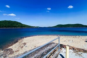A beach with clear water, a sandy shore, and distant green hills. - Nature Lover’s Dream with Expansive Tiered Deck Spaces in Garfield, Arkansas (Garfield)