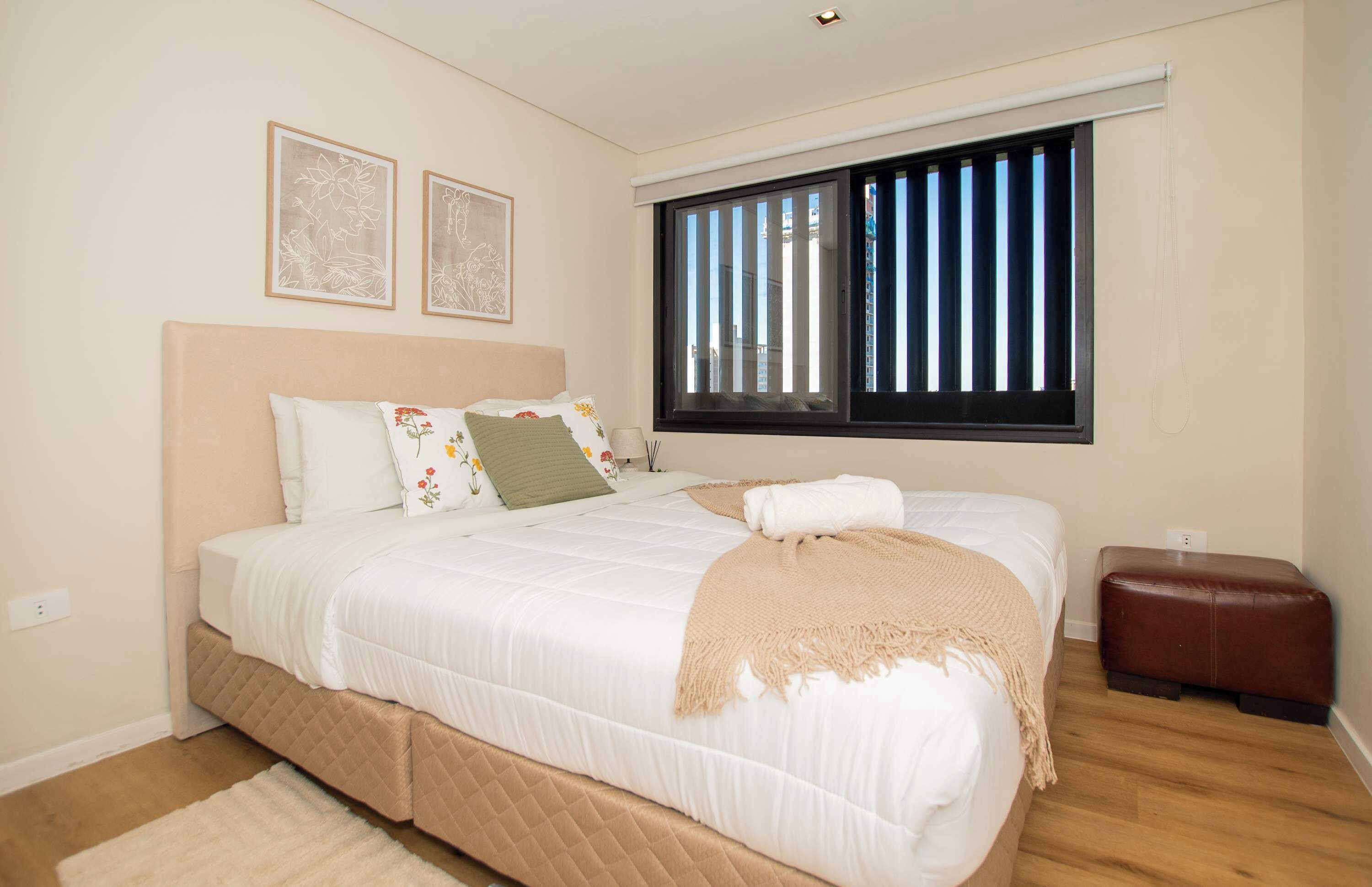Un dormitorio con cama, ventana con persianas, mesita de noche y enchufe en la pared.