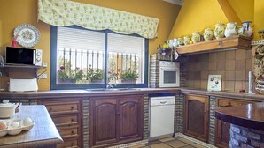 A kitchen with wooden cabinets, a built-in oven, a microwave, and a window with a view of flowers. - Charming and Spacious Family Villa - Cortijo Sierra y Lago (Arcos de la Frontera)