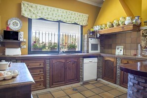A kitchen with wooden cabinets, a built-in oven, a microwave, and a window with a view of flowers. - Charming and Spacious Family Villa - Cortijo Sierra y Lago (Arcos de la Frontera)