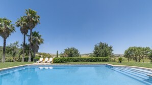 A rectangular swimming pool with a tiled deck, surrounded by palm trees and a hedge, with a clear blue sky in the background. - Charming and Spacious Family Villa - Cortijo Sierra y Lago (Arcos de la Frontera)