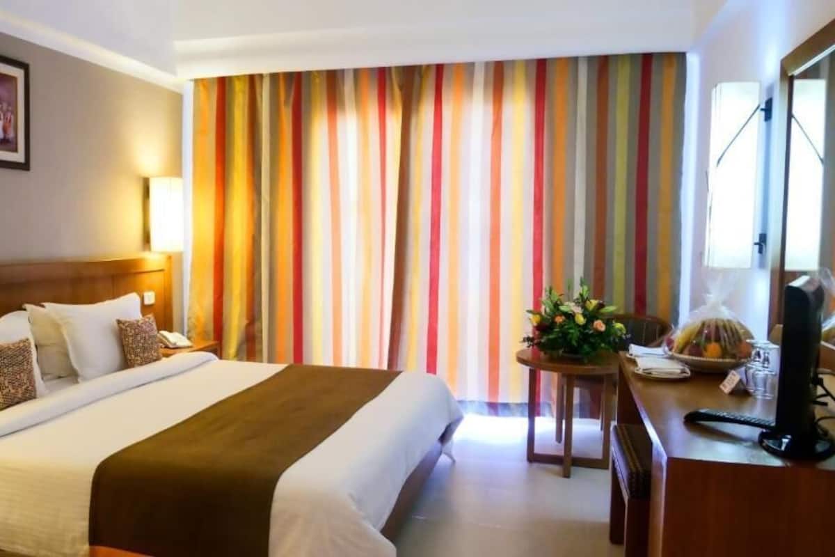 A hotel room with a bed, a desk, a chair, a lamp, a picture on the wall, and a window with curtains.