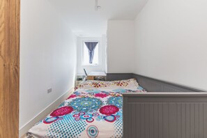 Ein schmaler Flur führt zu einem Schlafzimmer mit einem Bett, einem Fenster mit Vorhängen und einer weißen Tür.