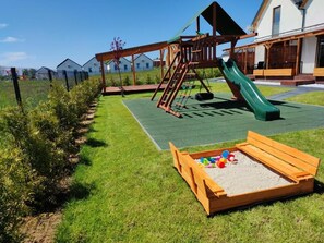 Ein Spielplatz mit Rutsche und Klettergerüst, umgeben von einem eingezäunten Bereich mit Rasen und Sandkasten.