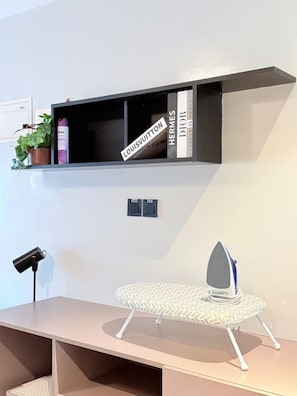 Une chambre moderne équipée d’un bureau, d’une planche à repasser, d’une étagère où sont exposés des articles de marques de luxe, et d’une plante en pot.