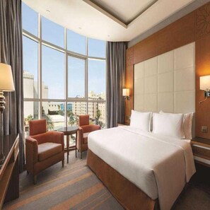 Une chambre d’hôtel avec un grand lit, deux fauteuils, une petite table et une vue sur le paysage urbain grâce à de grandes fenêtres.