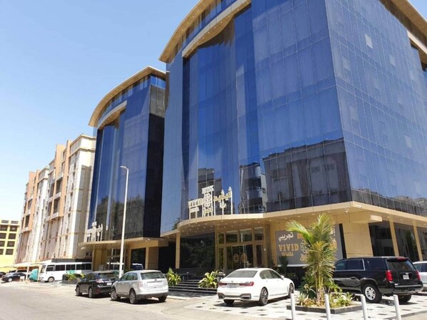 Un bâtiment moderne doté d’une façade en verre réfléchissant, d’une entrée imposante avec une enseigne et d’un espace de stationnement où sont garées plusieurs voitures.