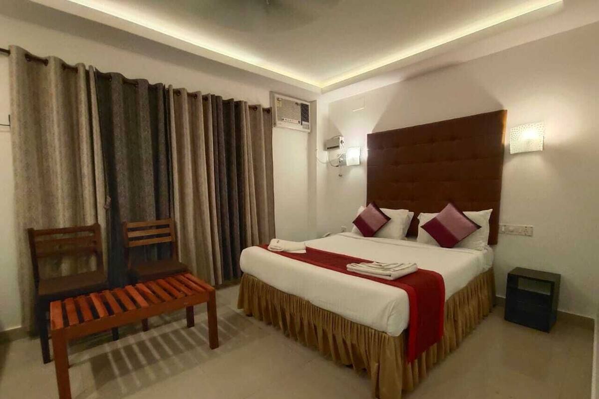 A hotel room with a bed, a bench, a bedside table, and a wall-mounted air conditioner.